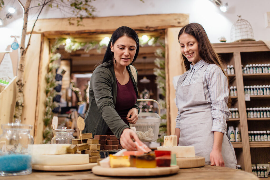 cheerful shopkeeper helping customer in packaging free shop. zero waste shopping woman choosing natural homemade soap at package free store.