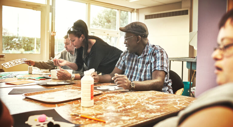 artistic, creative people with learning disabilities in class at a community center. a black woman is teaching the group while the adults learn, listen and enjoy arts and crafts together.